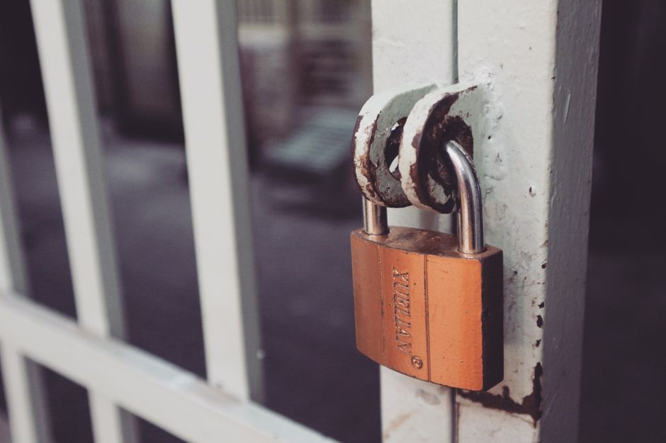 A close-up of a rusty, weathered metal padlock attached to a white-painted metal gate, with chipped paint revealing the underlying metal. The padlock is orange-brown and secured through the gate's lat